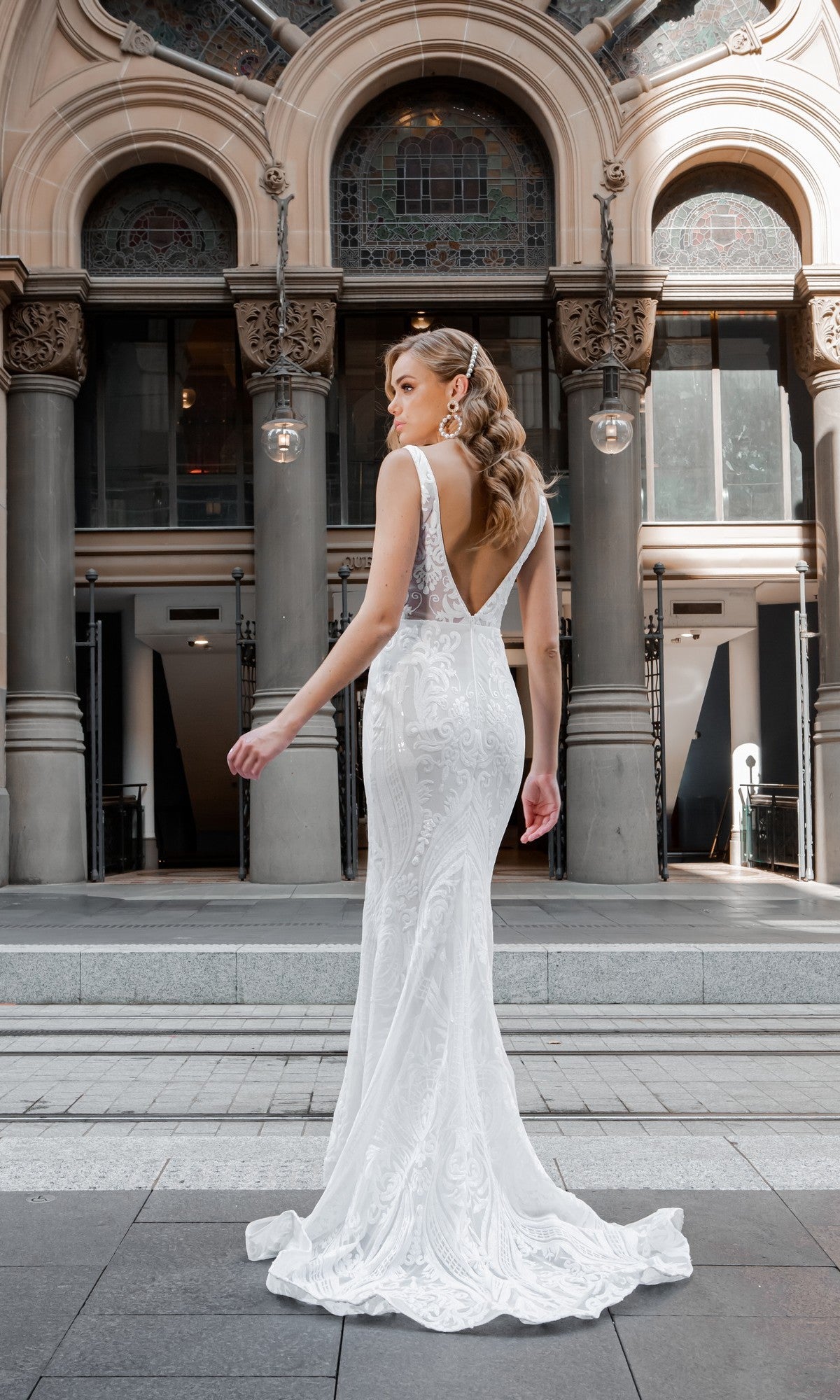 A woman in the Tina Holly BA109 White Lace Mermaid Wedding Gown stands outdoors before a building with columns and arched windows.